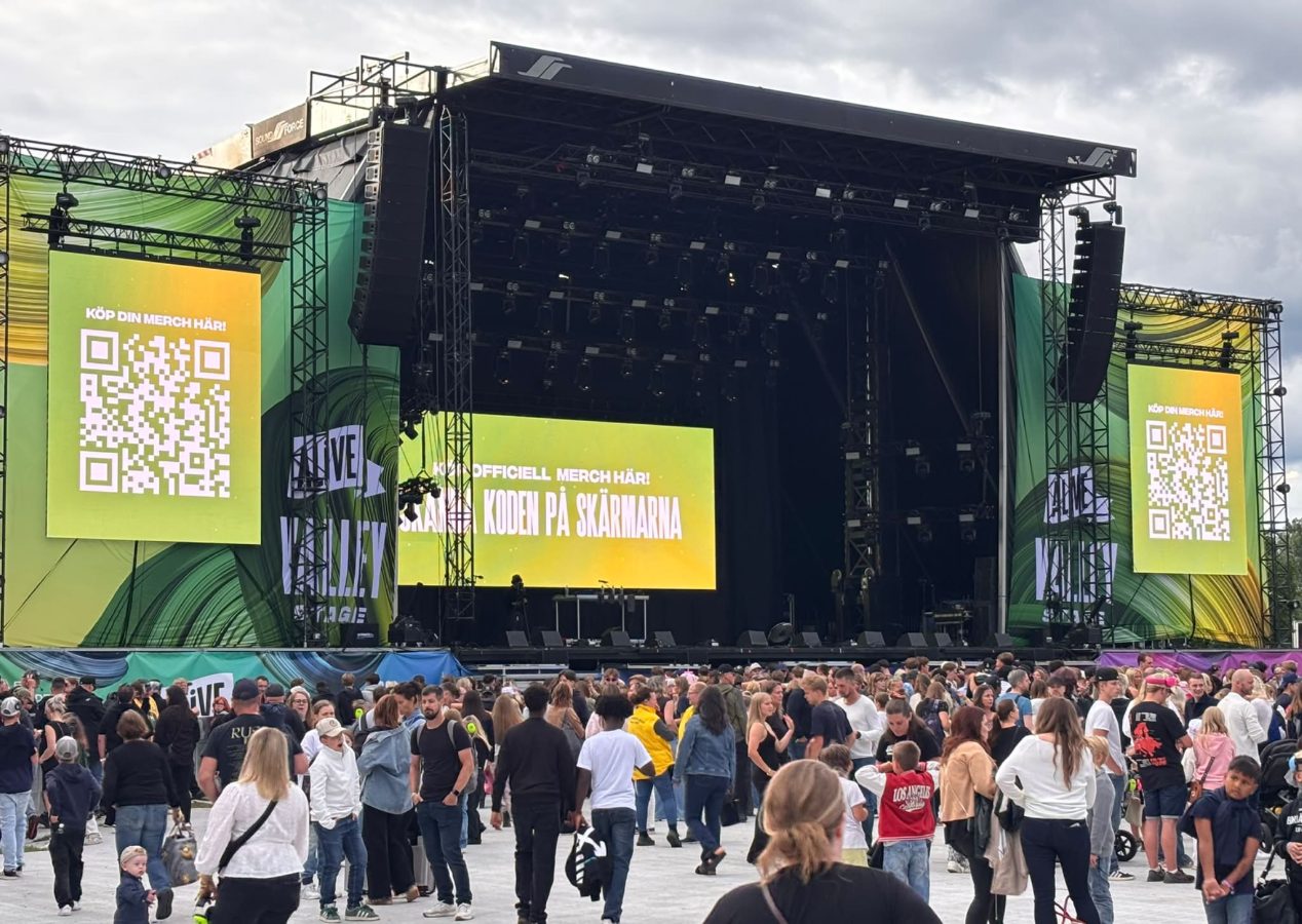 Storbildsskärmar med QR-koder och texten "Köp din merch här!" vid Alive Valley Stage.